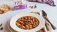 Ciotola di pasta e fagioli con pancetta Levoni servita calda con un piatto di ceramica bianco su tavolo elegante, con rosmarino fresco, filo d'olio extravergine e pane casereccio sullo sfondo. 