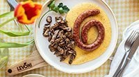 Polenta cremosa con Luganega Levoni adagiata sopra la polenta e accompagnata da funghi misti, servita su un piatto da portata da condividere in un ambiente primaverile e rustico. 