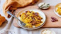 Spaghetti zucca cremosa e salsiccia, condita con Salsiccia Levoni e cubetti di zucca mantovana, servita in piatto bianco di ceramica, rametti di rosmarino e pepe nero, su una tovaglia chiara e canovaccio arancione. 