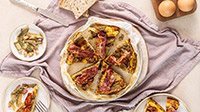 Frittata di carciofi alta e soffice con cubetti di Pancetta Levoni croccante e Bacon Levoni tostato come decorazione,  servita su piatto rustico con carciofi trifolati su sfondo di tavola in pietra naturale chiara e luce diffusa.