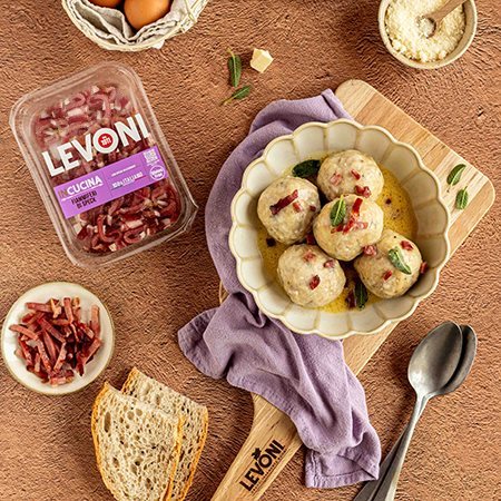 Canederli allo speck con Fiammiferi di Speck Levoni serviti con una cremina di burro e salvia, in un piatto fondo rustico appoggiato su un tagliere Levoni. 