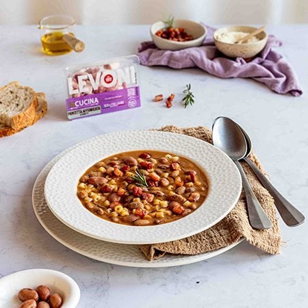 Ciotola di pasta e fagioli con pancetta Levoni servita calda con un piatto di ceramica bianco su tavolo elegante, con rosmarino fresco, filo d'olio extravergine e pane casereccio sullo sfondo. 