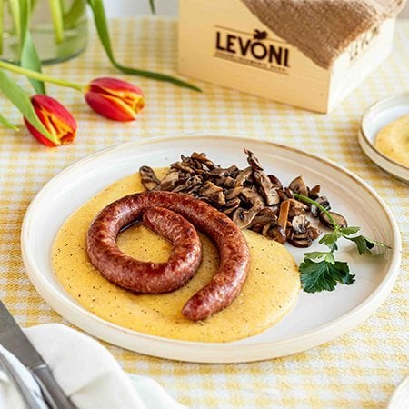 Polenta cremosa con Luganega Levoni adagiata sopra la polenta e accompagnata da funghi misti, servita su un piatto da portata da condividere in un ambiente primaverile e rustico. 