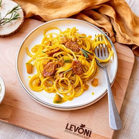 Spaghetti zucca cremosa e salsiccia, condita con Salsiccia Levoni e cubetti di zucca mantovana, servita in piatto bianco di ceramica, rametti di rosmarino e pepe nero, su una tovaglia chiara e canovaccio arancione. 