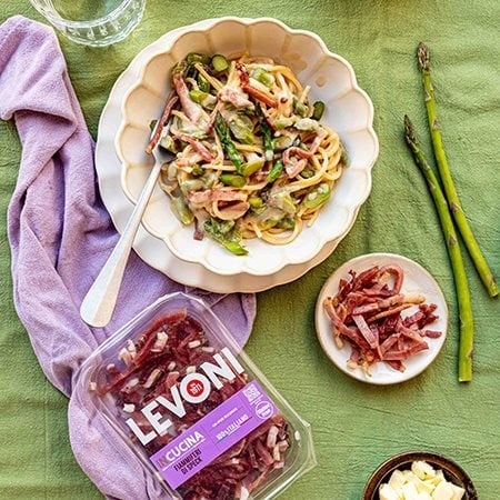 Pasta spaghetti con asparagi freschi e fiammiferi di Speck Levoni servita in piatto romantico e tovaglia verde con punte di asparago verdi in primo piano e tovagliolo rosa accanto. 
