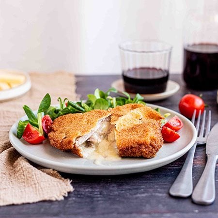 Cotoletta alla Valdostana con Prosciutto Cotto Alta Qualità Levoni e fontina filante, panatura dorata e croccante, servita su piatto in ceramica con guarnizione di insalata valeriana e pomodorini tagliati su tavola in legno scuro.  