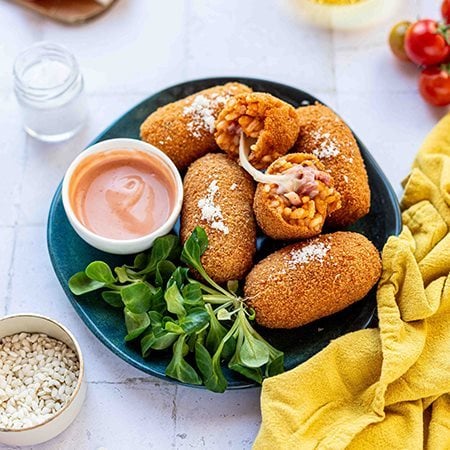 Supplì di riso con Prosciutto Cotto Alta Qualità Levoni e mozzarella filante, dorati e croccanti, serviti su piatto colorato con filo di mozzarella che si allunga al morso e una ciotolina di salsa rosa in una tavola moderna e contemporanea. 