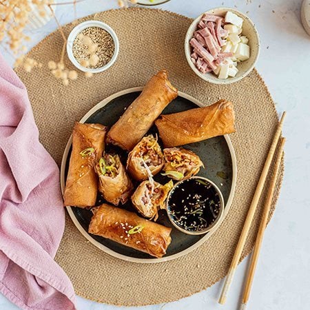 Involtini primavera all’italiana con Prosciutto Cotto Alta Qualità Levoni e verdure fresche, cotti al forno e dorati, serviti su un piatto nero in ceramica con salsa di soia aromatizzata con cipollotto e sesamo bianco accompagnato da bacchette orientali. 