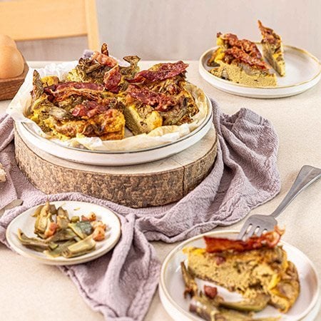 Frittata di carciofi alta e soffice con cubetti di Pancetta Levoni croccante e Bacon Levoni tostato come decorazione,  servita su piatto rustico con carciofi trifolati su sfondo di tavola in pietra naturale chiara e luce diffusa.