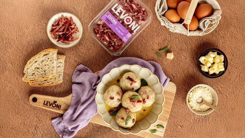 Canederli allo speck con Fiammiferi di Speck Levoni serviti con una cremina di burro e salvia, in un piatto fondo rustico appoggiato su un tagliere Levoni. 