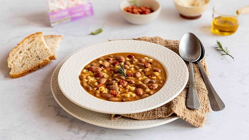 Ciotola di pasta e fagioli con pancetta Levoni servita calda con un piatto di ceramica bianco su tavolo elegante, con rosmarino fresco, filo d'olio extravergine e pane casereccio sullo sfondo. 