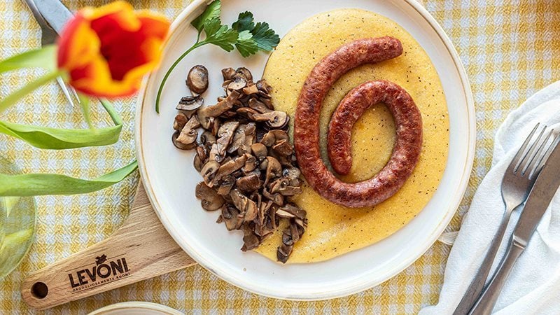 Polenta cremosa con Luganega Levoni adagiata sopra la polenta e accompagnata da funghi misti, servita su un piatto da portata da condividere in un ambiente primaverile e rustico. 