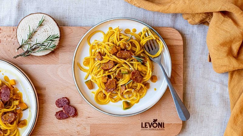 Spaghetti zucca cremosa e salsiccia, condita con Salsiccia Levoni e cubetti di zucca mantovana, servita in piatto bianco di ceramica, rametti di rosmarino e pepe nero, su una tovaglia chiara e canovaccio arancione. 