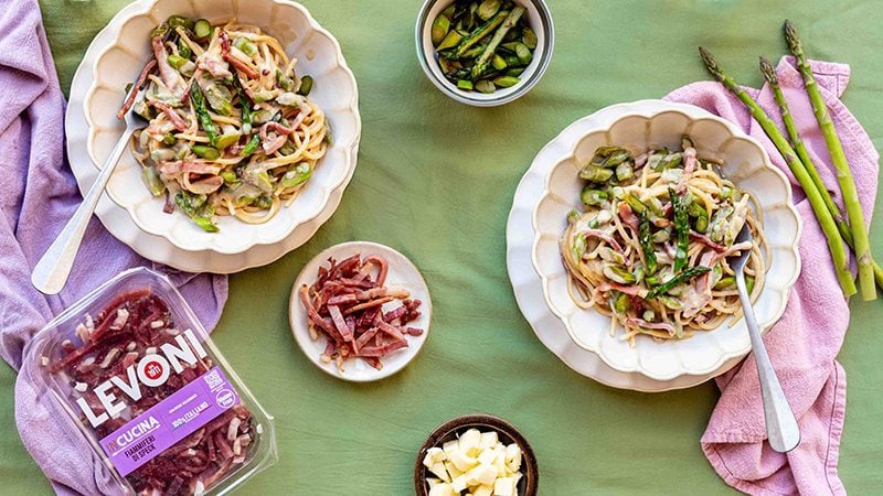 Pasta spaghetti con asparagi freschi e fiammiferi di Speck Levoni servita in piatto romantico e tovaglia verde con punte di asparago verdi in primo piano e tovagliolo rosa accanto. 