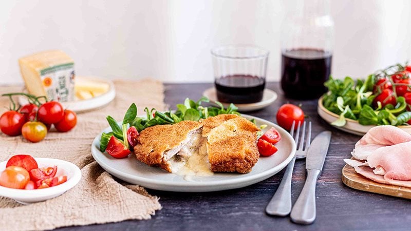 Cotoletta alla Valdostana con Prosciutto Cotto Alta Qualità Levoni e fontina filante, panatura dorata e croccante, servita su piatto in ceramica con guarnizione di insalata valeriana e pomodorini tagliati su tavola in legno scuro.  