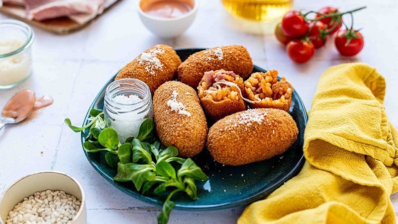 Supplì di riso con Prosciutto Cotto Alta Qualità Levoni e mozzarella filante, dorati e croccanti, serviti su piatto colorato con filo di mozzarella che si allunga al morso e una ciotolina di salsa rosa in una tavola moderna e contemporanea. 