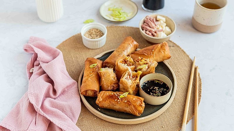 Involtini primavera all’italiana con Prosciutto Cotto Alta Qualità Levoni e verdure fresche, cotti al forno e dorati, serviti su un piatto nero in ceramica con salsa di soia aromatizzata con cipollotto e sesamo bianco accompagnato da bacchette orientali. 