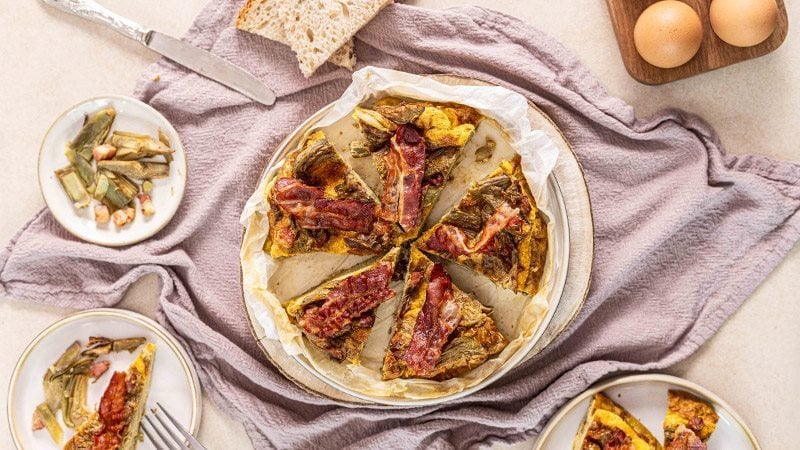 Frittata di carciofi alta e soffice con cubetti di Pancetta Levoni croccante e Bacon Levoni tostato come decorazione,  servita su piatto rustico con carciofi trifolati su sfondo di tavola in pietra naturale chiara e luce diffusa.