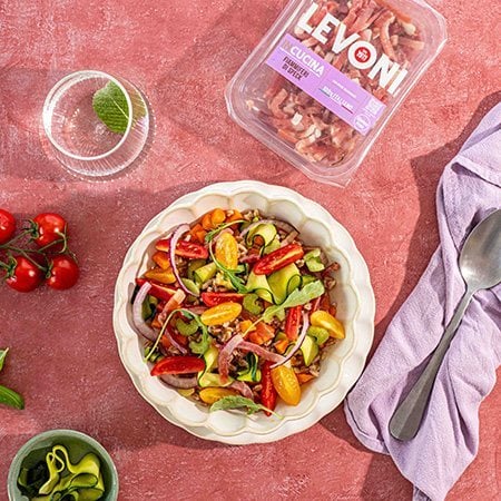 insalata di farro fredda con cubetti di Speck Levoni, pomodorini datterini tagliati a metà, dadini di zucchina e peperone, mandorle a lamelle e scaglie di grana, servita in una ciotola di ceramica chiara su un tavolo in legno con foglie di basilico fresco sparse intorno. 