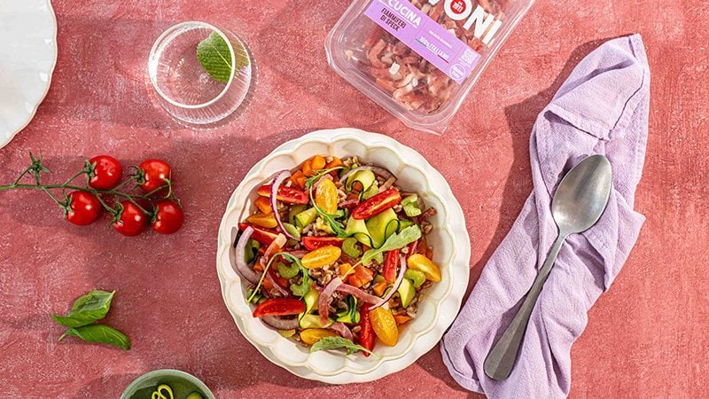 insalata di farro fredda con cubetti di Speck Levoni, pomodorini datterini tagliati a metà, dadini di zucchina e peperone, mandorle a lamelle e scaglie di grana, servita in una ciotola di ceramica chiara su un tavolo in legno con foglie di basilico fresco sparse intorno. 