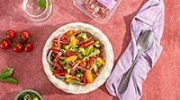 insalata di farro fredda con cubetti di Speck Levoni, pomodorini datterini tagliati a metà, dadini di zucchina e peperone, mandorle a lamelle e scaglie di grana, servita in una ciotola di ceramica chiara su un tavolo in legno con foglie di basilico fresco sparse intorno. 