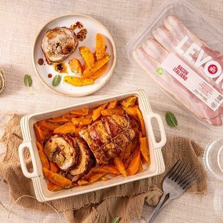 Arrosto di vitello avvolto nel Bacon italiano Levoni, dorato e appoggiato su una teglia in ceramica con fondo di cottura lucido, rametti di rosmarino fresco e carote al prezzemolo in spicchi, servito su tavola imbandita per il pranzo della domenica.