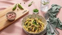 Fusilli con piselli freschi e Prosciutto Cotto Alta Qualità Levoni, cremosa e profumata, servita in piatto fondo romantico sopra una tavola allestita con una tovaglia primaverile, un tagliere e ingredienti della ricetta. 