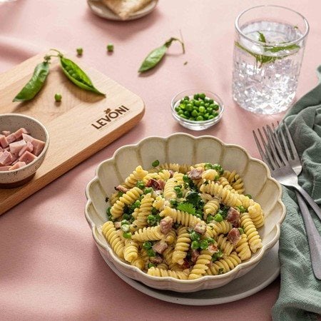 Fusilli con piselli freschi e Prosciutto Cotto Alta Qualità Levoni, cremosa e profumata, servita in piatto fondo romantico sopra una tavola allestita con una tovaglia primaverile, un tagliere e ingredienti della ricetta. 