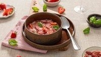 Risotto fragole e Fiammiferi di Speck Levoni cremoso e dal colore rosa intenso, servito in una ciotola piana con fragole fresche intere, listarelle di speck croccante e foglioline di basilico fresco, sopra una tovaglia neutra e un tagliare di legno chiaro con fragole sparse.