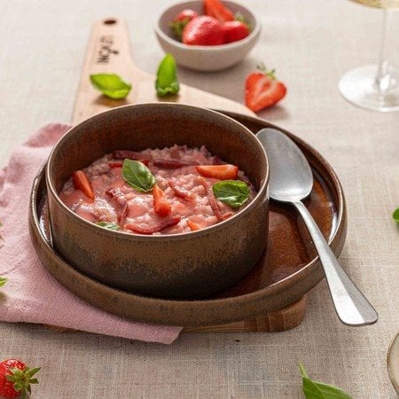 Risotto fragole e Fiammiferi di Speck Levoni cremoso e dal colore rosa intenso, servito in una ciotola piana con fragole fresche intere, listarelle di speck croccante e foglioline di basilico fresco, sopra una tovaglia neutra e un tagliare di legno chiaro con fragole sparse.