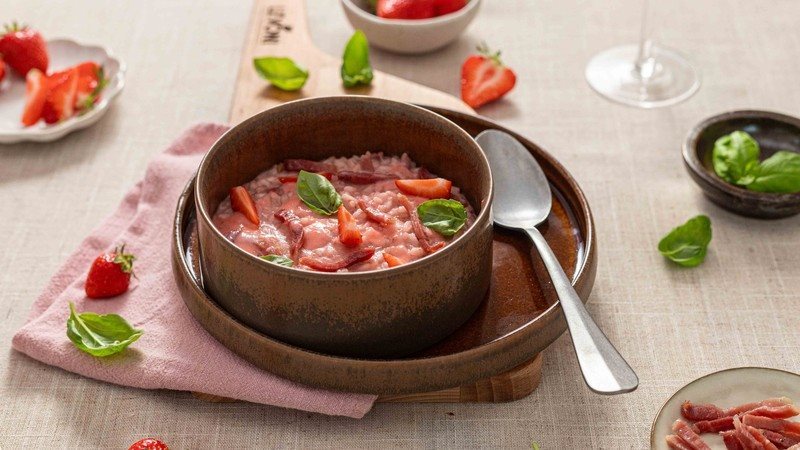 Risotto fragole e Fiammiferi di Speck Levoni cremoso e dal colore rosa intenso, servito in una ciotola piana con fragole fresche intere, listarelle di speck croccante e foglioline di basilico fresco, sopra una tovaglia neutra e un tagliare di legno chiaro con fragole sparse.