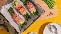 Asparagi verdi gratinati al forno con Speck Alto Adige Levoni e fontina valdostana filante, serviti in teglia con speck croccante, punte di asparago e fili di formaggio che si sciolgono, su sfondo di tavola primaverile e colorata. 