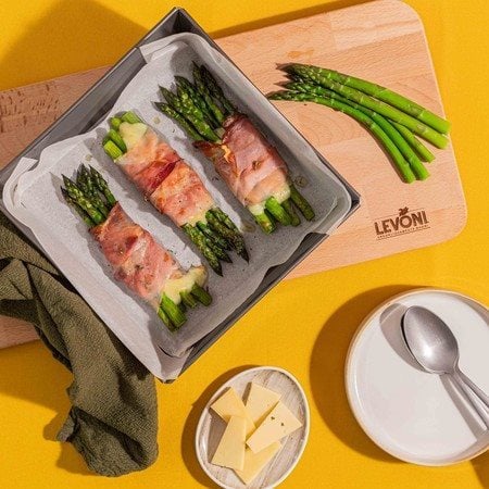 Asparagi verdi gratinati al forno con Speck Alto Adige Levoni e fontina valdostana filante, serviti in teglia con speck croccante, punte di asparago e fili di formaggio che si sciolgono, su sfondo di tavola primaverile e colorata. 