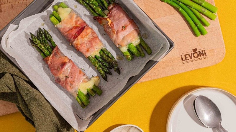 Asparagi verdi gratinati al forno con Speck Alto Adige Levoni e fontina valdostana filante, serviti in teglia con speck croccante, punte di asparago e fili di formaggio che si sciolgono, su sfondo di tavola primaverile e colorata. 