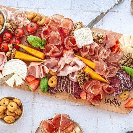 Tagliere di salumi e formaggi con Prosciutto Crudo, Speck, Mortadella, pancetta arrotolata e salami misti Levoni, formaggi misti a scaglie e a pasta molle, accompagnato da fragole, pomodorini ciliegini e melone, grissini artigianali, presentato su tagliere in legno rustico con foglie di basilico e ciotoline varie.