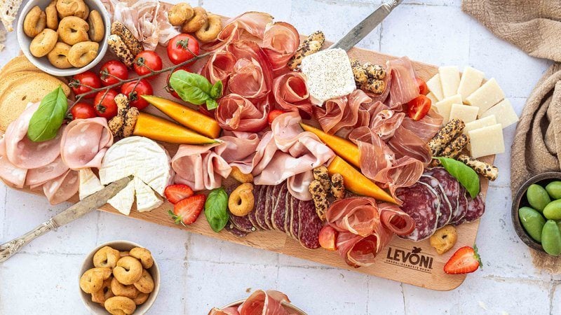 Tagliere di salumi e formaggi con Prosciutto Crudo, Speck, Mortadella, pancetta arrotolata e salami misti Levoni, formaggi misti a scaglie e a pasta molle, accompagnato da fragole, pomodorini ciliegini e melone, grissini artigianali, presentato su tagliere in legno rustico con foglie di basilico e ciotoline varie.