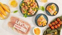 Antipasto di prosciutto crudo e melone con fette e palline di melone retato maturo e fette di Prosciutto Crudo Levoni adagiate su piatto in ceramica scura con della valeriana come accompagnamento, in stile mediterraneo estivo.