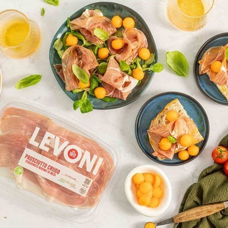 Antipasto di prosciutto crudo e melone con fette e palline di melone retato maturo e fette di Prosciutto Crudo Levoni adagiate su piatto in ceramica scura con della valeriana come accompagnamento, in stile mediterraneo estivo.