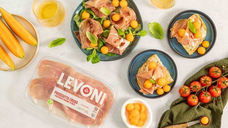 Antipasto di prosciutto crudo e melone con fette e palline di melone retato maturo e fette di Prosciutto Crudo Levoni adagiate su piatto in ceramica scura con della valeriana come accompagnamento, in stile mediterraneo estivo.