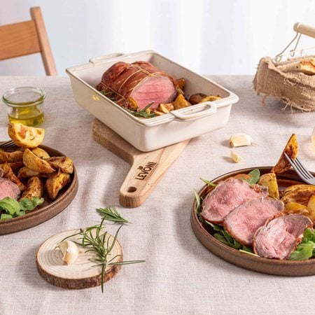 roast beef con crosta di Speck Levoni affettato al centro, con il cuore rosato ben visibile, adagiato all’interno di una pirofila con patate arrosto in spicchi e rametti di rosmarino fresco, appoggiato sopra un tagliere e su una tavola imbandita con elementi naturali. 