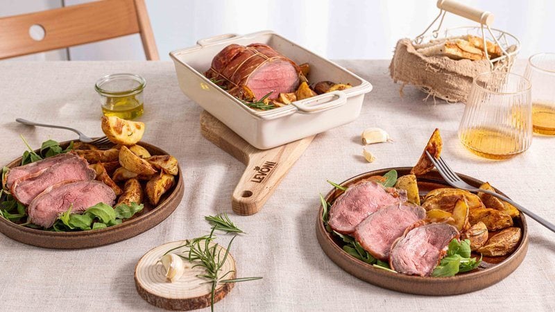 roast beef con crosta di Speck Levoni affettato al centro, con il cuore rosato ben visibile, adagiato all’interno di una pirofila con patate arrosto in spicchi e rametti di rosmarino fresco, appoggiato sopra un tagliere e su una tavola imbandita con elementi naturali. 