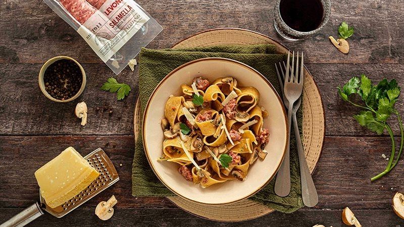 piatto di pasta alla boscaiola con salsiccia Levoni, funghi e panna, servito in piatto di ceramica bianca con prezzemolo fresco e spolverata di Parmigiano, illuminato da luce naturale.