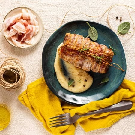 filetto di maiale avvolto in Pancetta Levoni, dorato e croccante, affettato e servito adagiato su un purè di patate e insaporito da rosmarino e salvia fresco su piatto piano. 