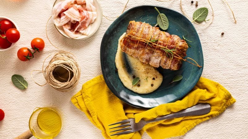 filetto di maiale avvolto in Pancetta Levoni, dorato e croccante, affettato e servito adagiato su un purè di patate e insaporito da rosmarino e salvia fresco su piatto piano. 